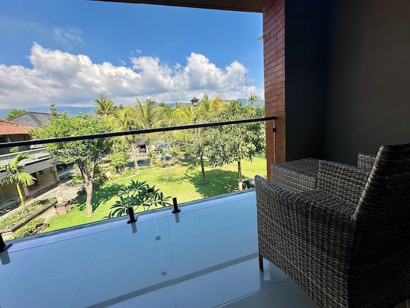 A balcony with a wicker chair, a view of a garden with palm trees, and a clear sky.
