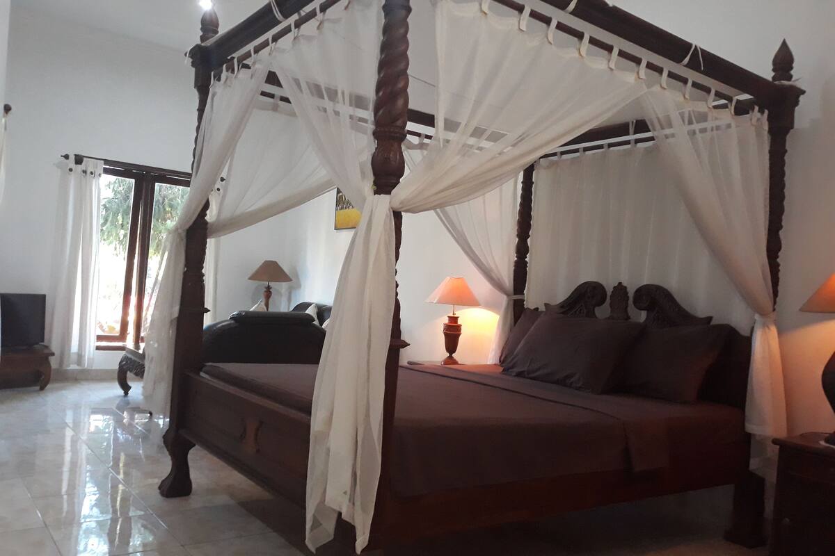 A four-poster canopy bed with a wooden frame and white drapes, accompanied by two bedside lamps and a nightstand.