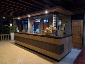 Un mostrador de recepción con un letrero que indica la zona de recepción y caja, un arreglo decorativo y una planta en maceta.
