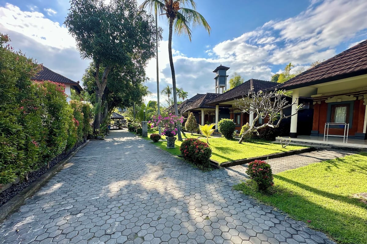 A pathway leading to a building with a tiled roof, surrounded by well-maintained greenery and a clear sky.