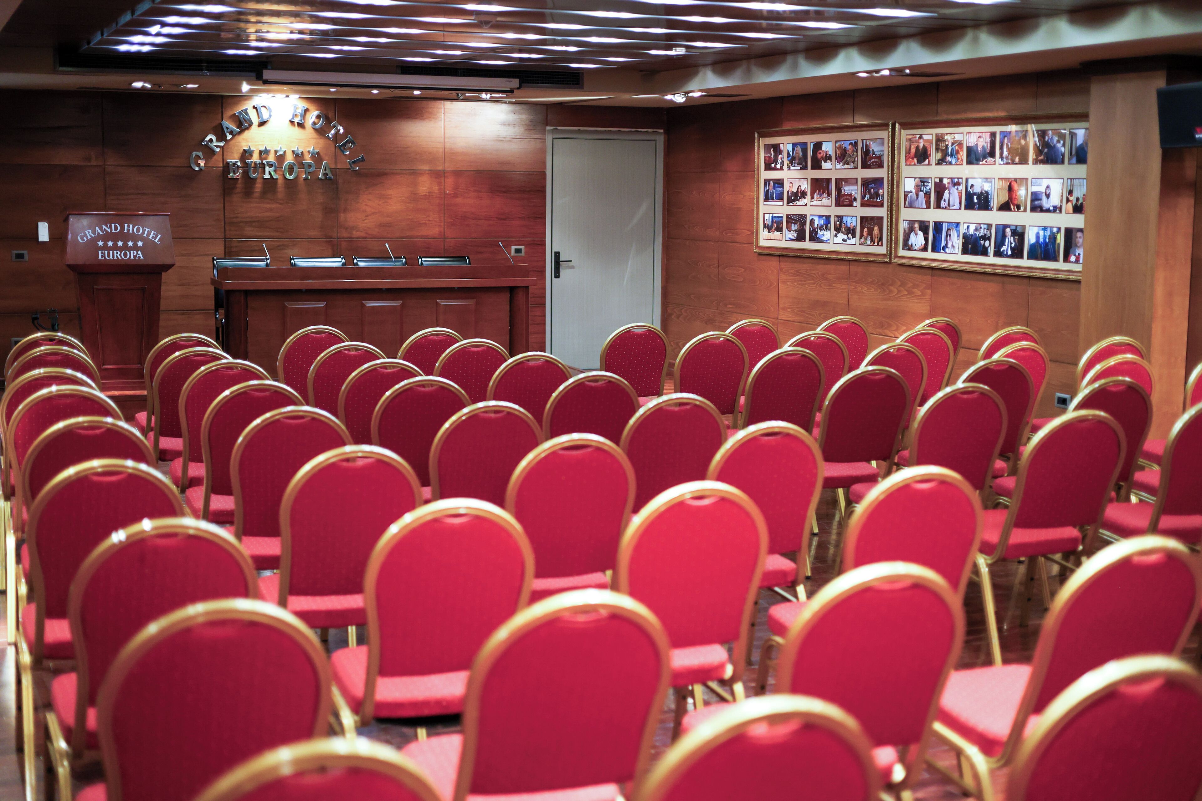 A conference room with rows of red chairs, a podium, and a wall with framed photos.