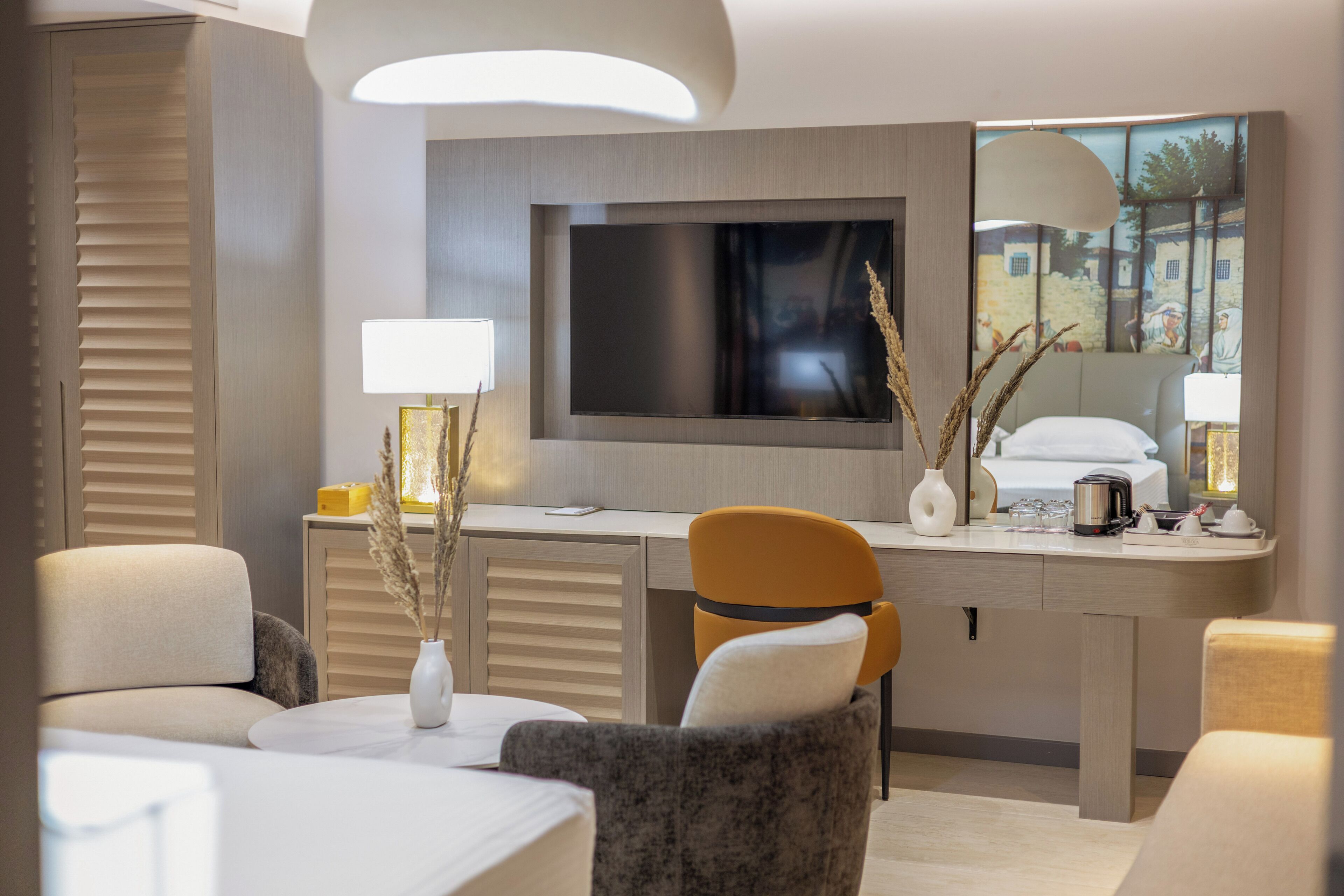 Quarto de hotel moderno com televisão de tela plana na parede, escrivaninha com luminária e área de estar confortável com cadeiras.