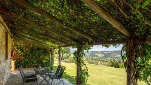 Un espace extérieur abrité, doté d’un sol en pierre, d’une table et de chaises, offrant une vue sur un paysage verdoyant avec des collines à l’horizon.