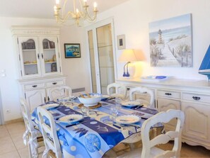 Un coin repas avec une table dressée pour six personnes, des chaises blanches, un lustre, un tableau représentant un phare et un buffet aux portes vitrées.