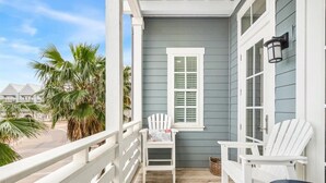 A porch with white chairs and a table, a window with shutters, and a view of palm trees and houses.
