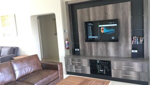 A modern living room with a large flat-screen TV mounted on a wooden wall, a leather sofa, a wooden coffee table, and built-in storage cabinets. - Caribbean Estates Villa & Apt (Port Edward)