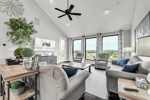A spacious living room with a ceiling fan, large windows, a TV, and a view of the outdoors.
