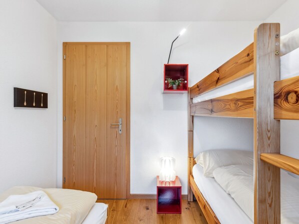 Une petite pièce avec une porte en bois, une petite table de chevet avec une lampe, un lit superposé et une étagère rouge ornée d’une plante.