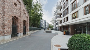 A pathway flanked by a brick building and modern apartment buildings, with a bench and greenery along the path. - Wooden Studio with Balcony | Parking | Close to Station (Kraków)