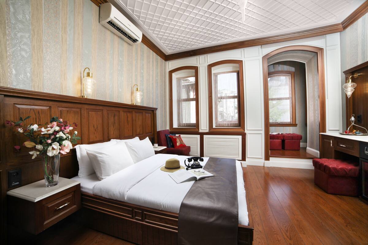 A hotel room with a large bed, wooden headboard, a nightstand with a vase of flowers, a desk with a lamp, and a view of the city through arched windows.