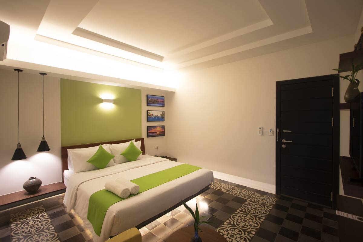 A hotel room with a bed, a nightstand, a lamp, a vase, and a door.