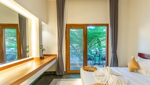 A hotel room with a large bed, a wooden bedside table, a mirror, and a view of the outdoors through a sliding glass door. - Baahu Villa (Siem Reap)