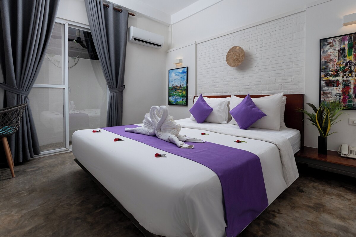 A hotel room with a large bed, a window with curtains, a painting on the wall, and a potted plant.