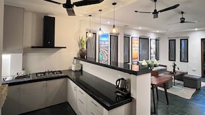 A modern kitchen with black countertops, white cabinets, and a central island. There are ceiling fans, pendant lights, and a large window with a cityscape view. - Baahu Villa (Siem Reap)
