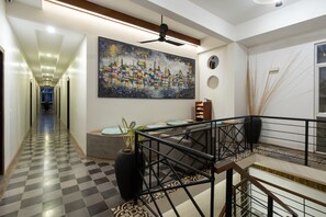 Un pasillo de hotel con piso a cuadros, un cuadro abstracto grande en la pared, un ventilador de techo y una escalera con barandilla.
