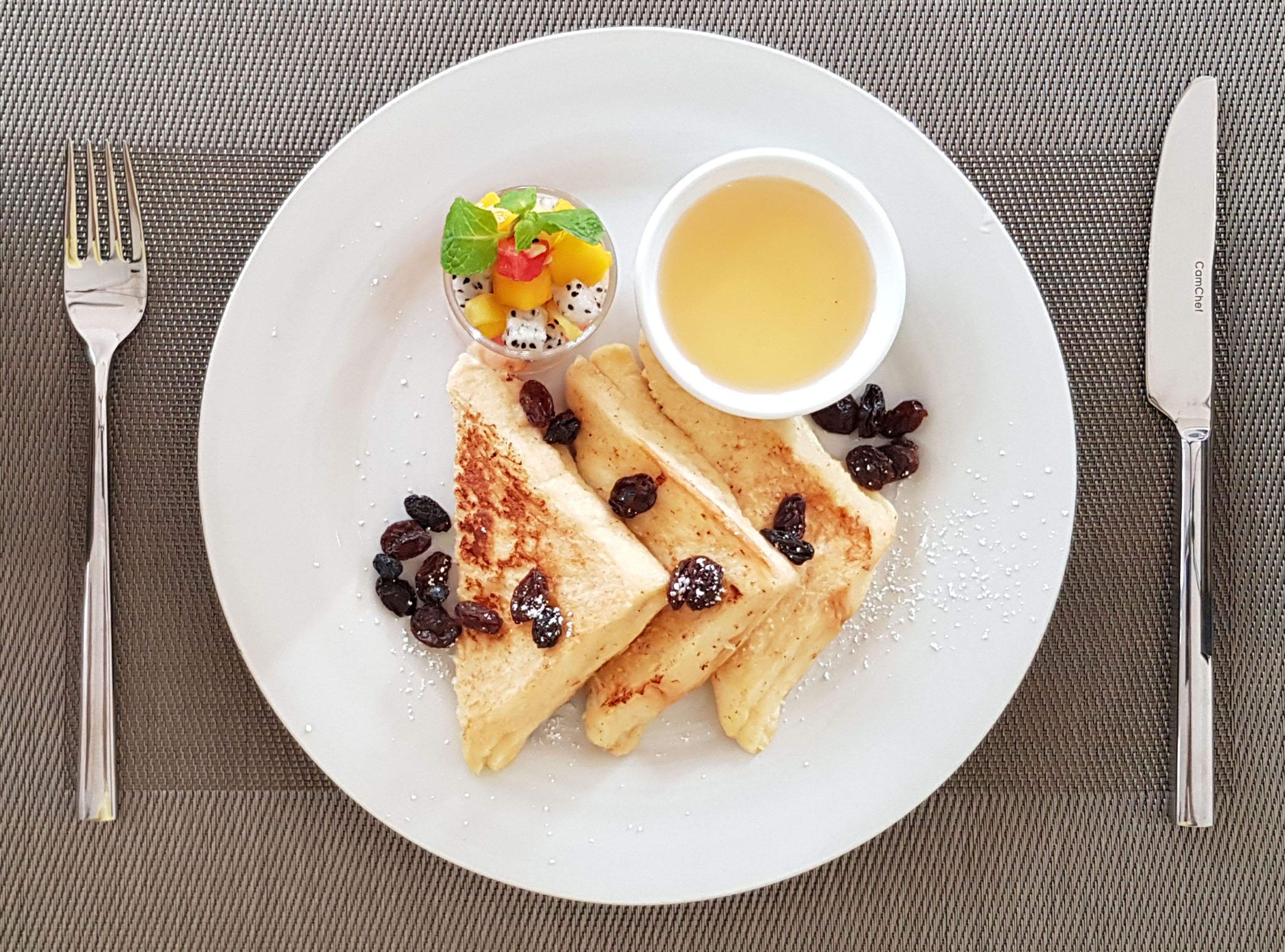 A plate with French toast, a fruit salad, and a cup of syrup.