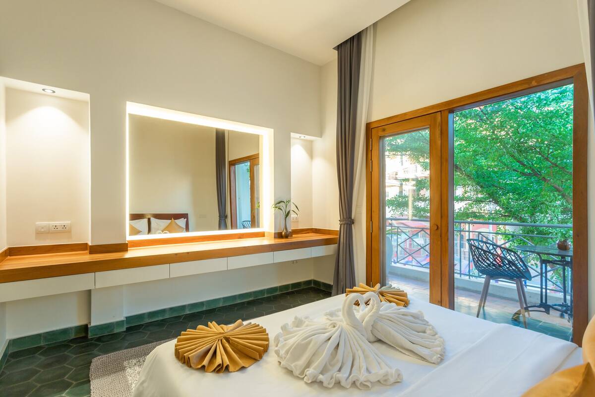 A hotel room with a large bed, a vanity with a mirror, a window with a view of greenery, and a balcony with outdoor furniture.
