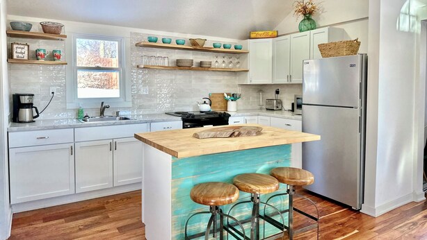 A modern kitchen with white cabinets, a stainless steel refrigerator, a wooden island with stools, and a window providing natural light. - Sunny Cottage: Fire pit, close to biking & walking trails and historic downtown. (Oskaloosa)