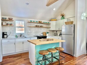 Cozinha moderna com armários brancos, geladeira de aço inoxidável, ilha de madeira com banquetas e uma janela que oferece iluminação natural.
