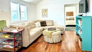 A living room with a sofa, ottoman, wooden floor, bookshelf, and a television. - Sunny Cottage: Fire pit, close to biking & walking trails and historic downtown. (Oskaloosa)