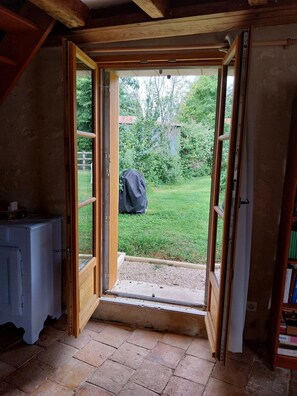 Une porte ouverte donnant sur un jardin avec une pelouse et des arbres, un objet recouvert d’une bâche et un sol carrelé.
