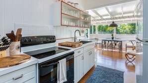 Una cocina moderna con armarios blancos, una estufa negra y una tabla de cortar de madera. Hay un comedor con mesa y sillas, y un ventanal con vistas al exterior.