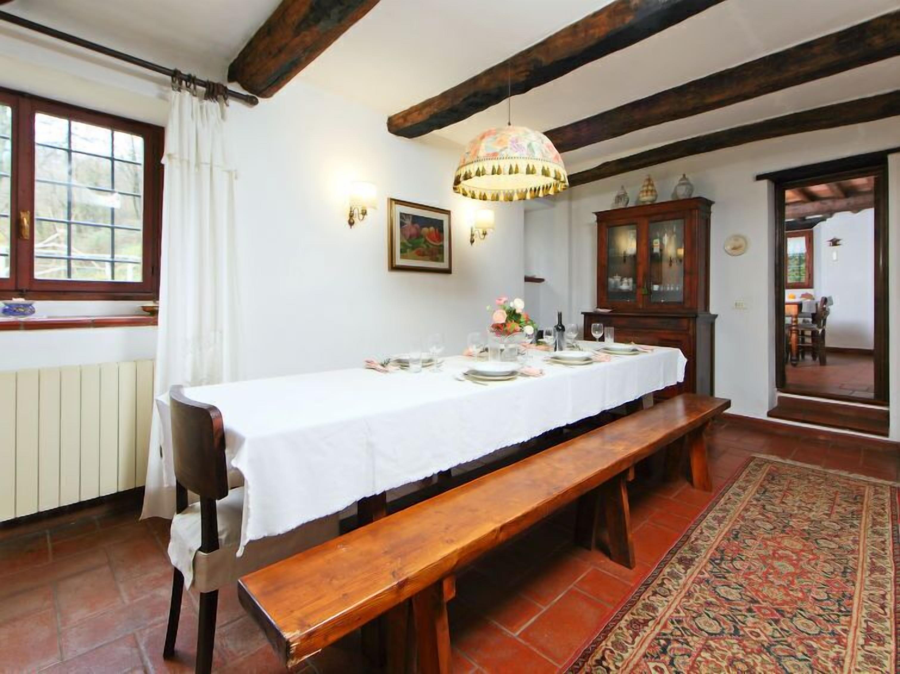 Un espace repas avec une longue table en bois dressée pour le repas, un banc, un tapis à motifs et un lustre.