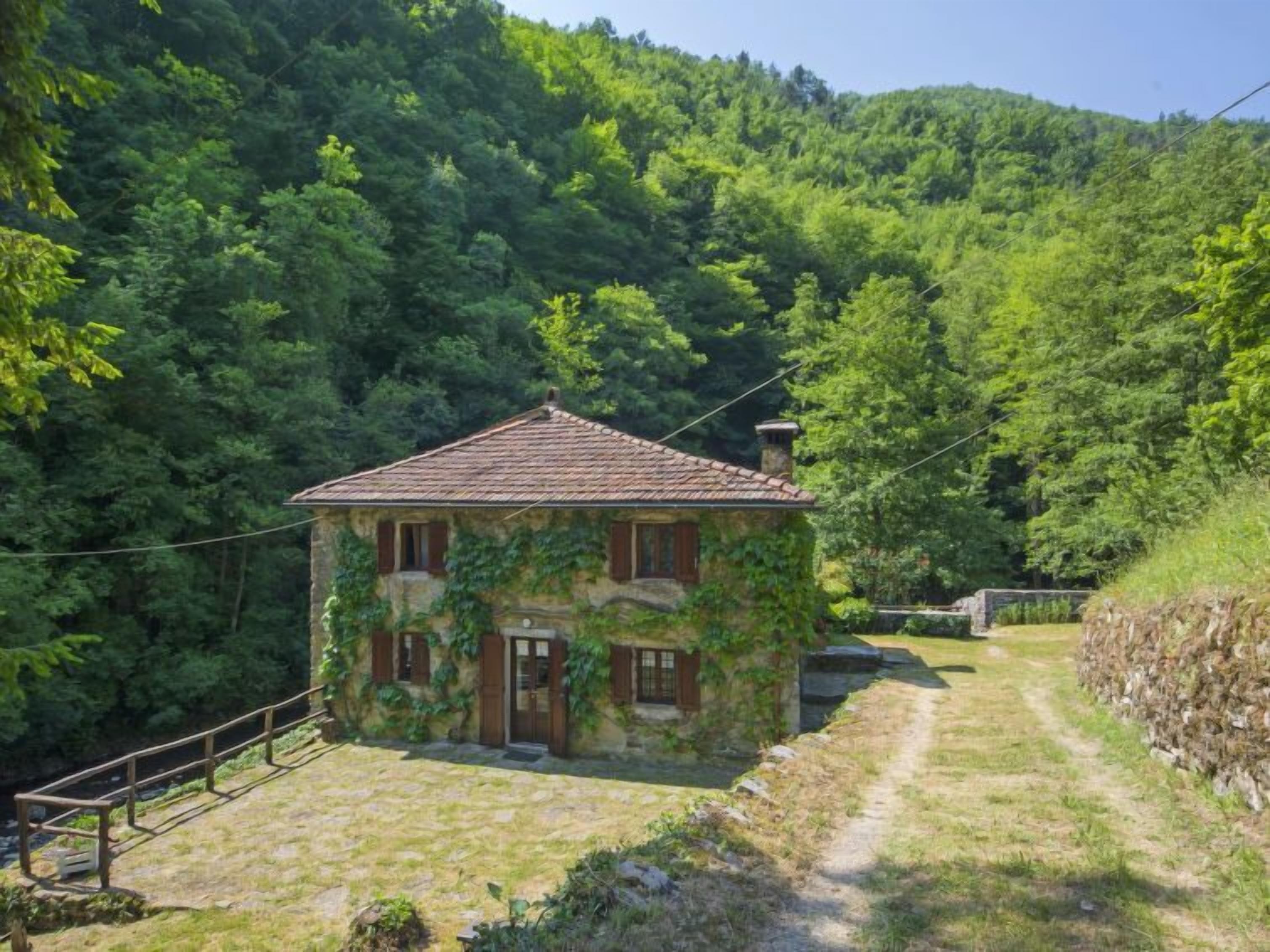 A stone house with a tiled roof, surrounded by lush greenery and a stone wall.