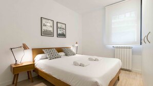 A neatly made bed with white linens and rolled towels, a wooden nightstand with a lamp, two framed pictures on the wall, and a window with a view of a building. - Gloria | The Rentals Collection (San Sebastián)