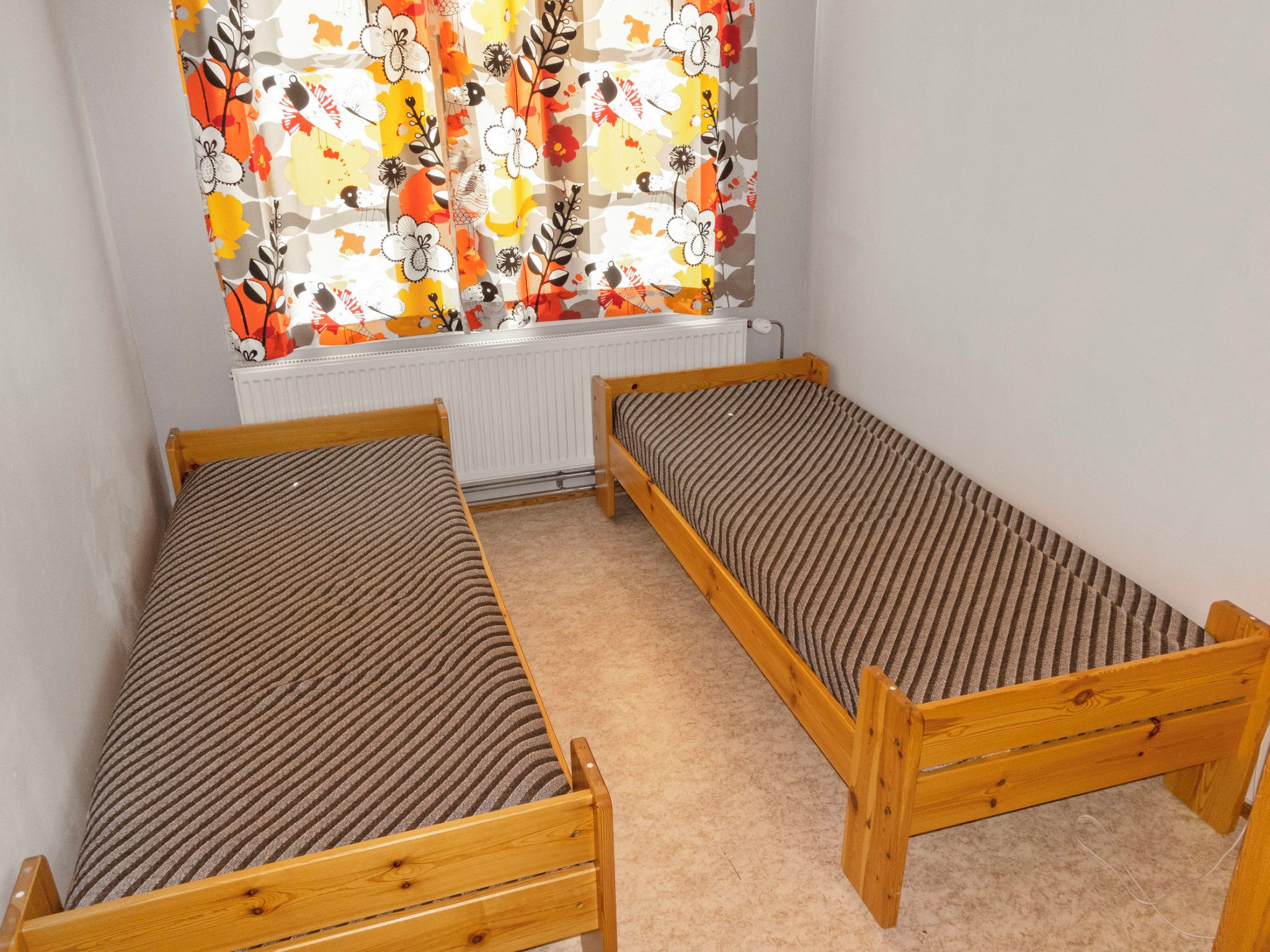 Ein Zimmer mit zwei Holzbettgestellen übereinander, einem Heizkörper und einem Fenster mit bunten Vorhängen.