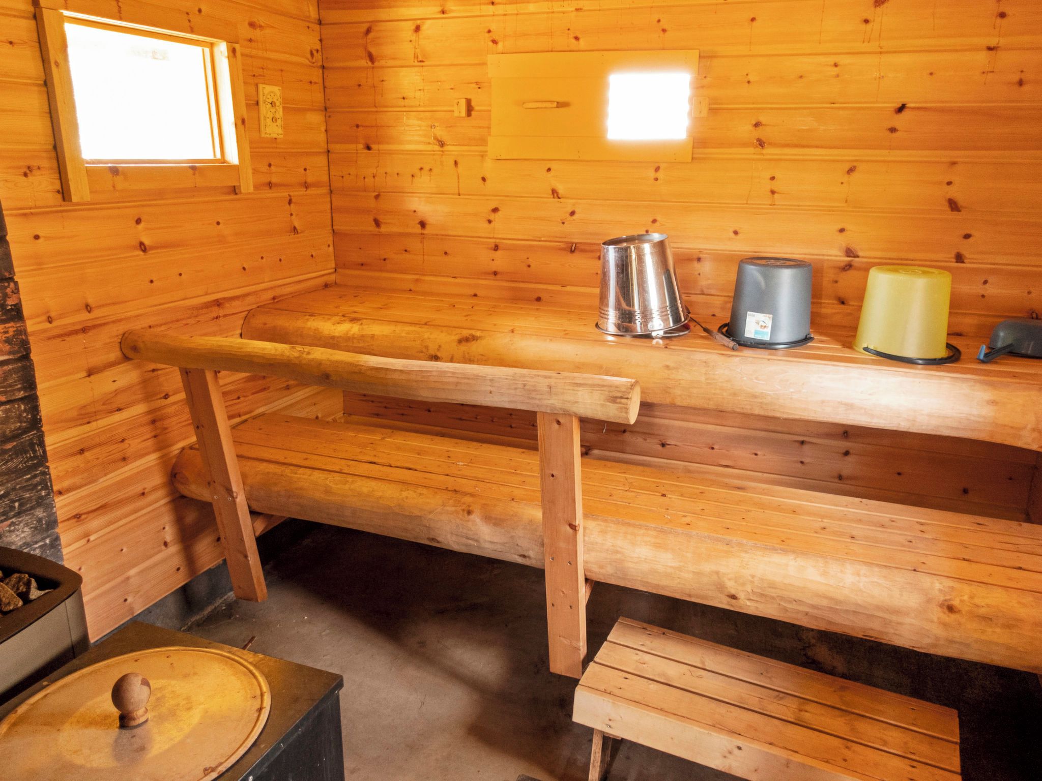 Interior de uma cabana de madeira com um banco, uma chaleira de metal e um recipiente amarelo numa prateleira.