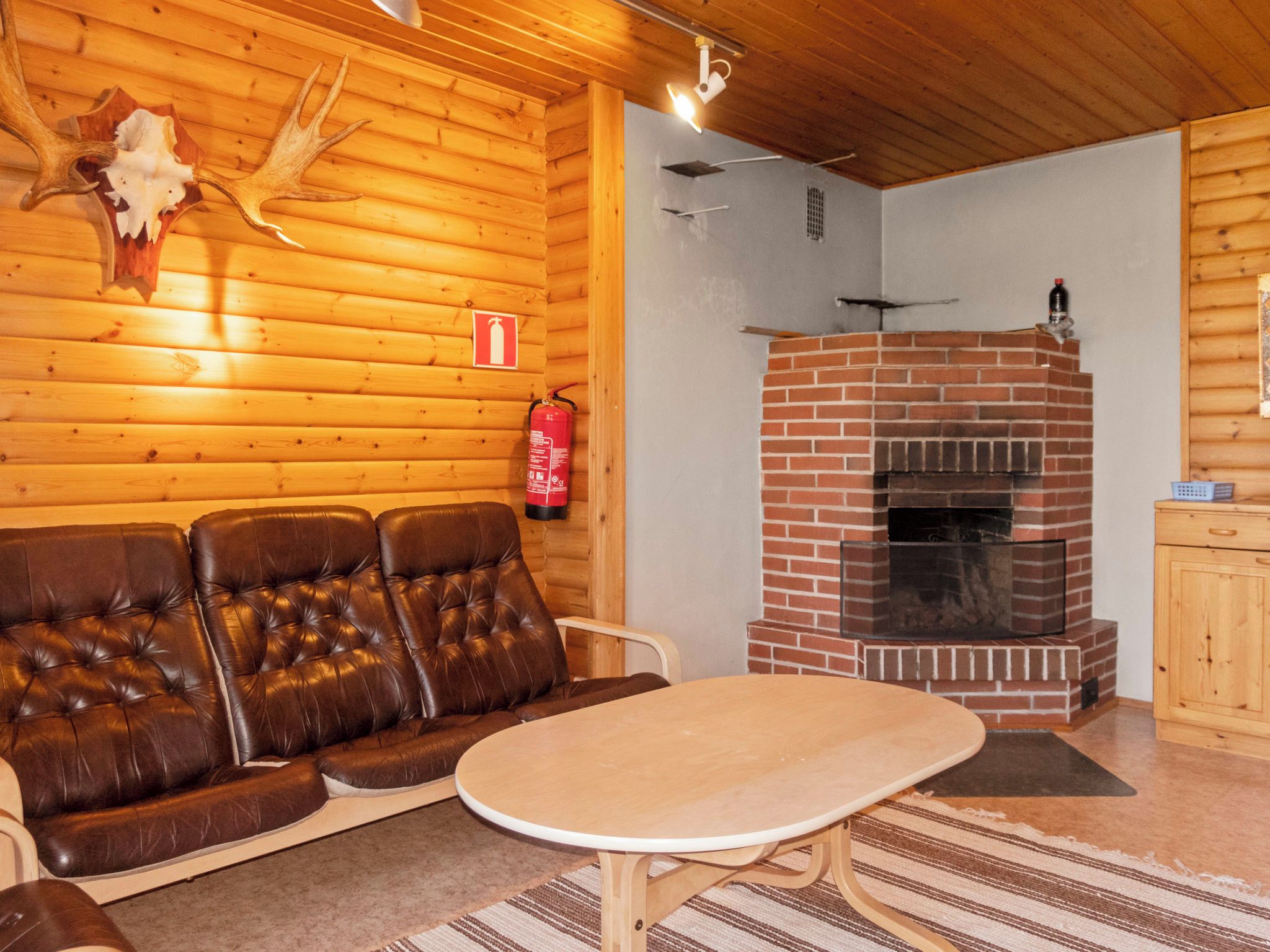 Interior de una cabaña de troncos con un sofá de cuero, una mesa redonda, una chimenea de ladrillo y un extintor de incendios montado en la pared.