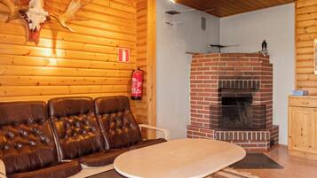 Interior de una cabaña de troncos con un sofá de cuero, una mesa redonda, una chimenea de ladrillo y un extintor de incendios montado en la pared.