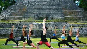Un grupo de personas practicando yoga sobre esterillas al aire libre, con antiguas estructuras de piedra como telón de fondo.