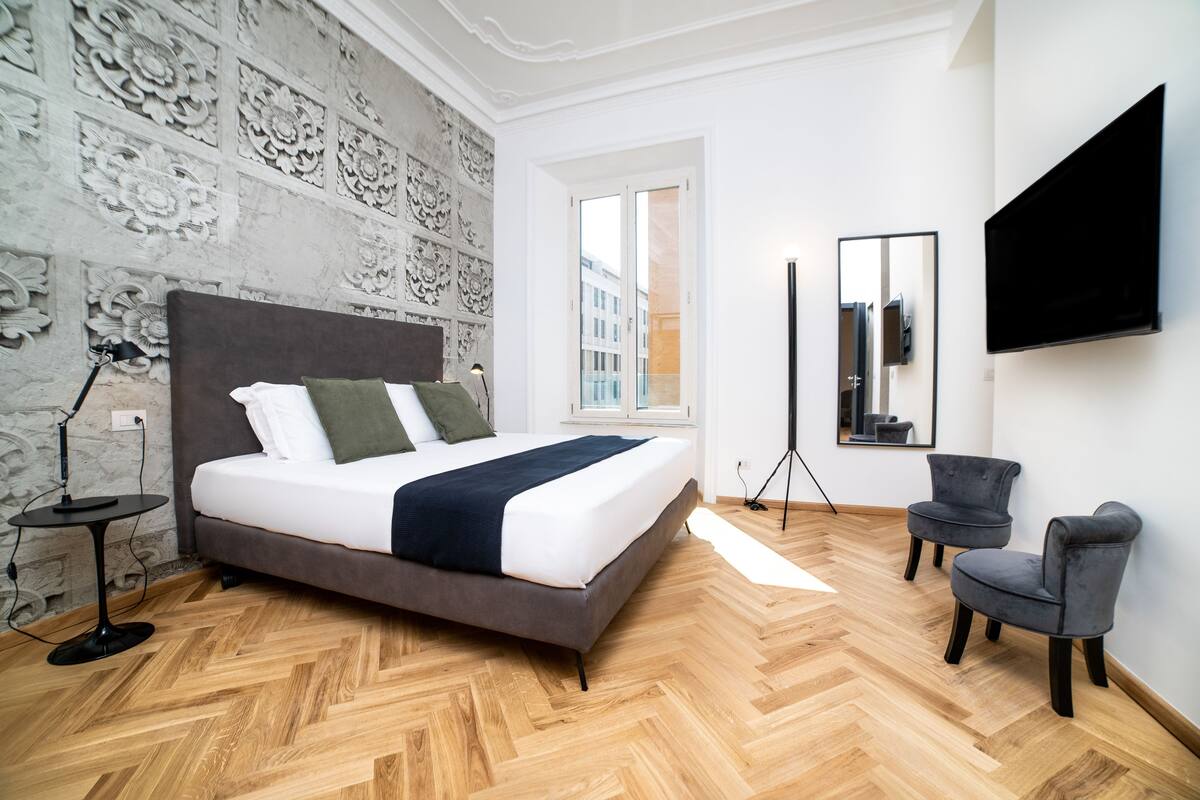 A modern bedroom with a large bed, a standing mirror, two armchairs, and a TV mounted on the wall.