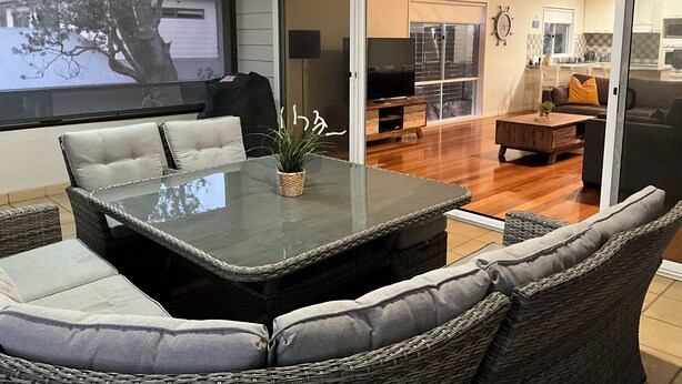 Una sala de estar moderna con una mesa de vidrio, muebles de mimbre y un gran ventanal corredera que da a una cocina.