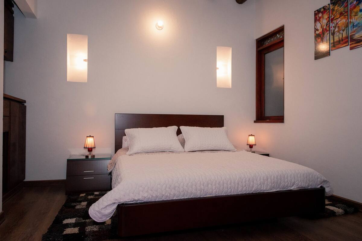 Un dormitorio con cabecera de madera, una cama grande con ropa de cama blanca, dos lámparas de noche, una mesita de noche y un espejo en la pared.