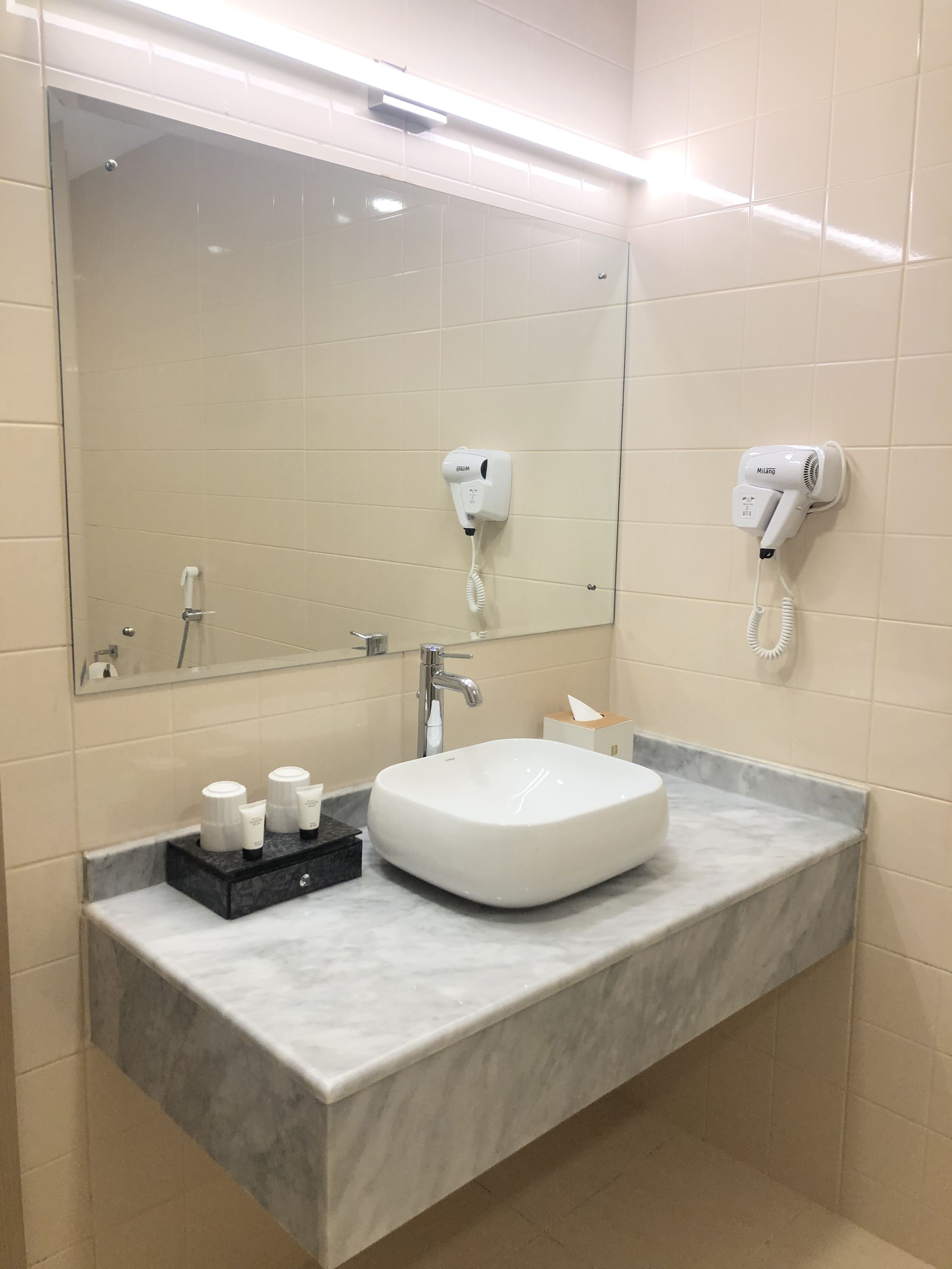A bathroom with a large mirror, a sink, a hairdryer, and a towel dispenser.