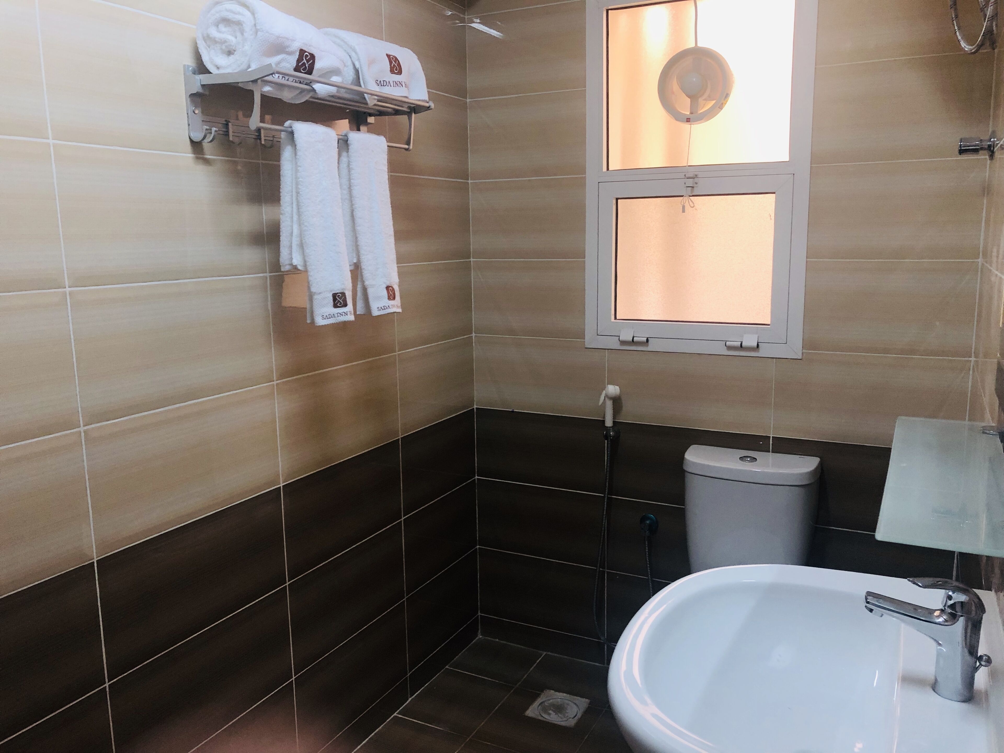 Baño con lavamanos, inodoro y ducha. Hay toallas colgadas en un perchero y una ventana con vistas.