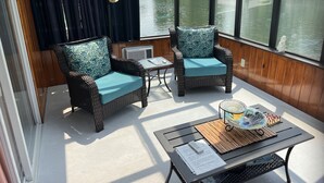 Ein gemütliches Zimmer mit Korbstühlen und einem Tisch, große Fenster mit Blick auf Wasser und Grünflächen.
