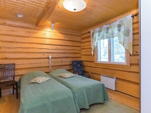 A wooden cabin room with two beds, a chair, a window with curtains, and a ceiling light.