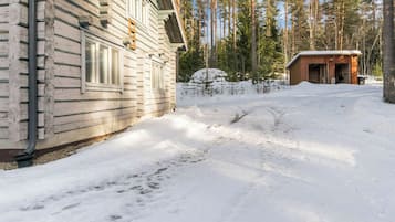 Una cabaña de troncos con un patio cubierto de nieve, un cobertizo de madera y un bosque al fondo.