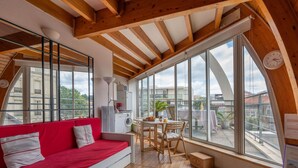 A cozy room with a red sofa, a dining table, and large windows offering a view of the city. - Apartment Hunier by Interhome (Arcachon)