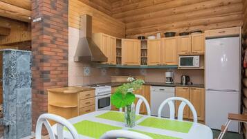 A wooden cabin interior with a kitchen, dining table, chairs, and a fireplace.