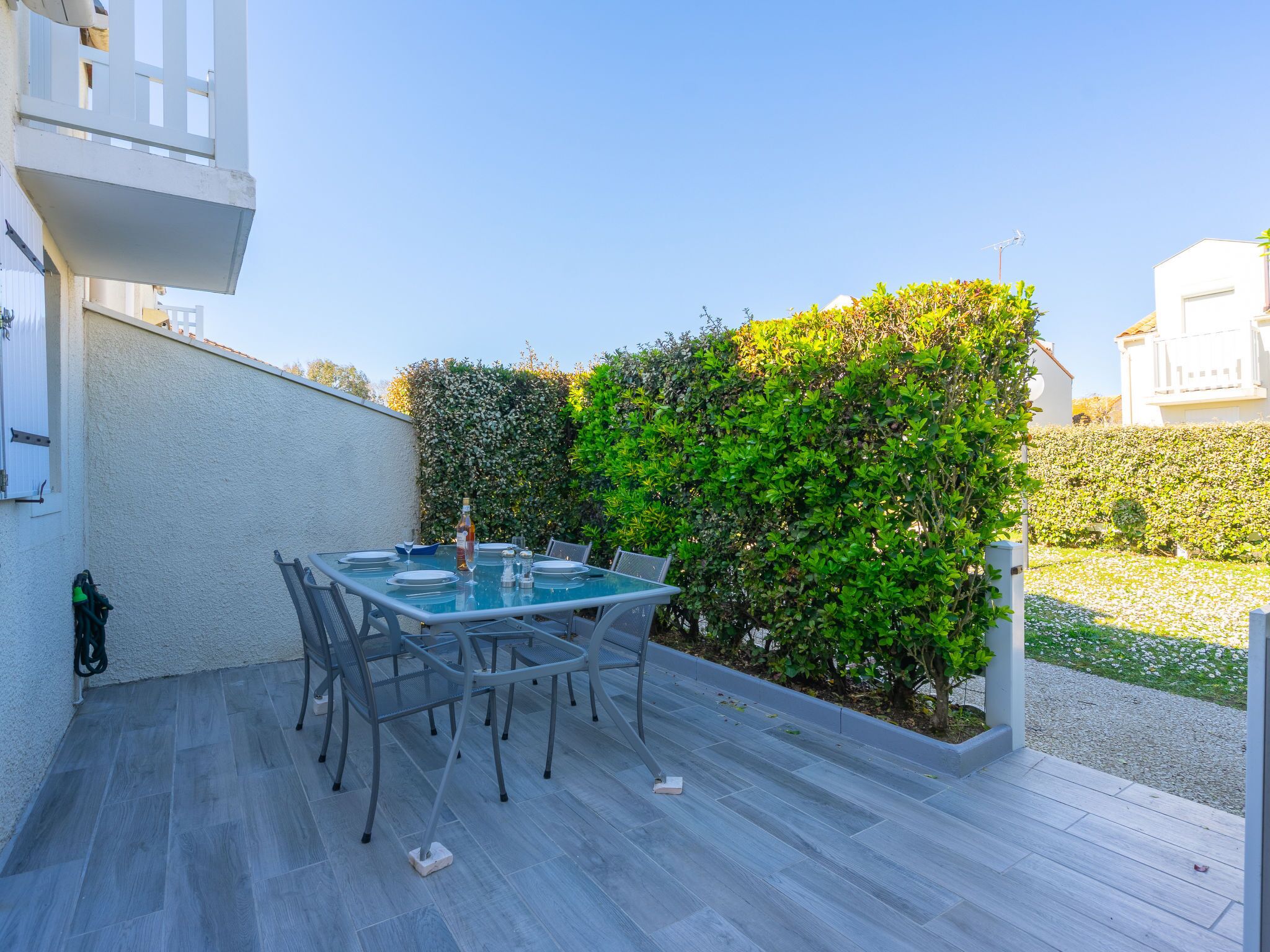 An outdoor patio with a glass table and chairs, surrounded by a hedge and a white wall.