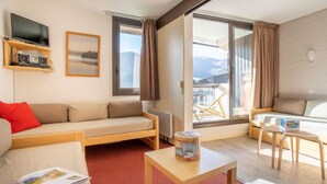 Una habitación de hotel moderna con un sofá, una mesita con una revista, un televisor, una radio y un balcón con vistas a las montañas.
