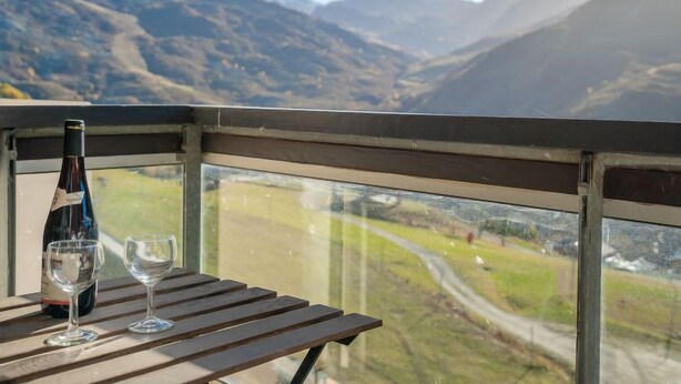 Un balcón con una mesa, una botella de vino y dos copas. Se ven montañas y una carretera.