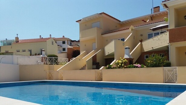 A swimming pool with a clear blue water, surrounded by a beige building with a red-tiled roof, and a staircase leading to a balcony with potted plants. - Wonderful villa composed of 3 bedrooms with wardrobes, 2 complete bathrooms. (Foz do Arelho)
