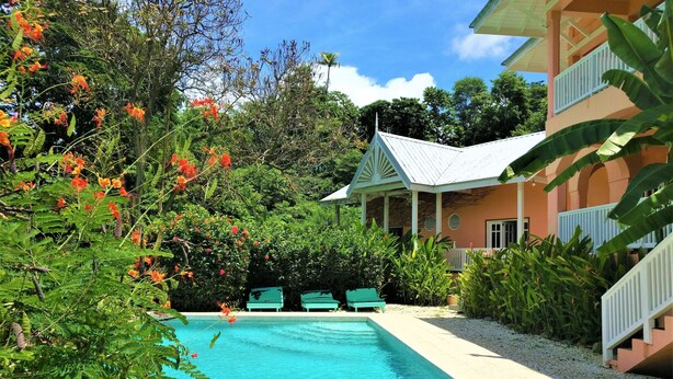A pool area with a clear blue pool, surrounded by lush greenery and a building with a white balcony. - Tobago Hibiscus Villas and Apartments (Bethel)