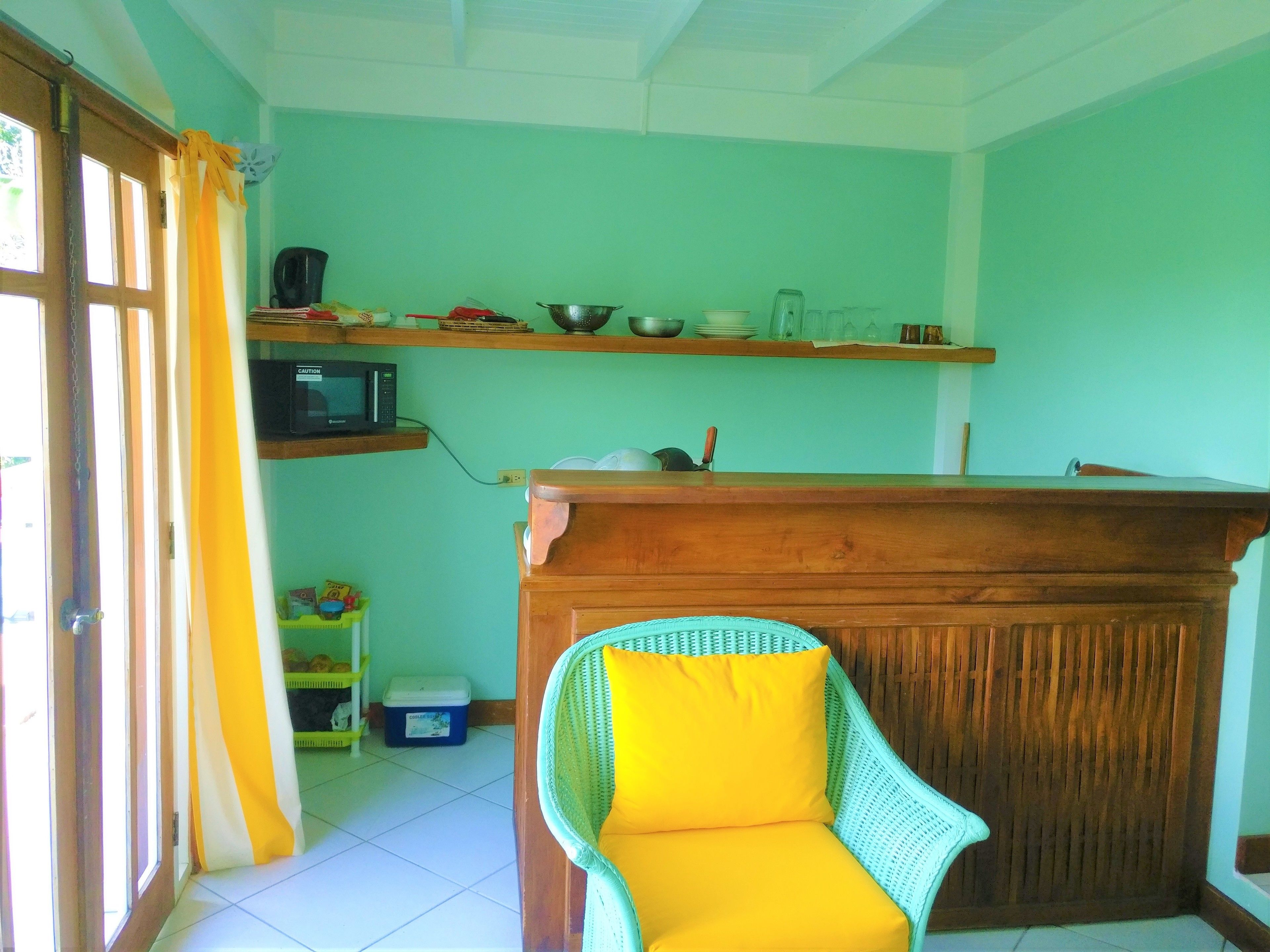 A room with a wooden reception desk, a wicker chair with a yellow cushion, a microwave on a shelf, and a window with curtains.